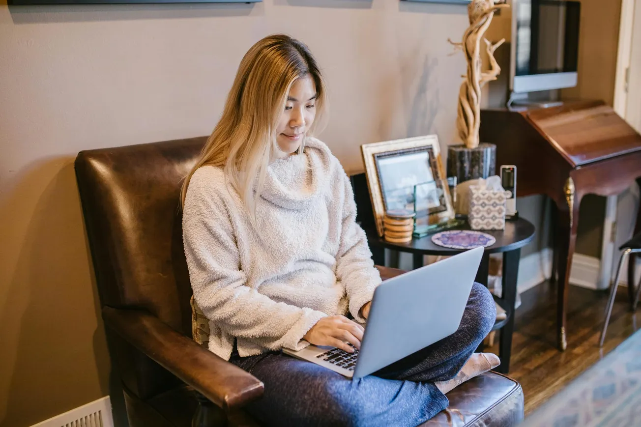 A woman in a cozy sweater sits cross-legged on a chair using a laptop. The room is warmly decorated with framed photos and a wooden sculpture on a side table.