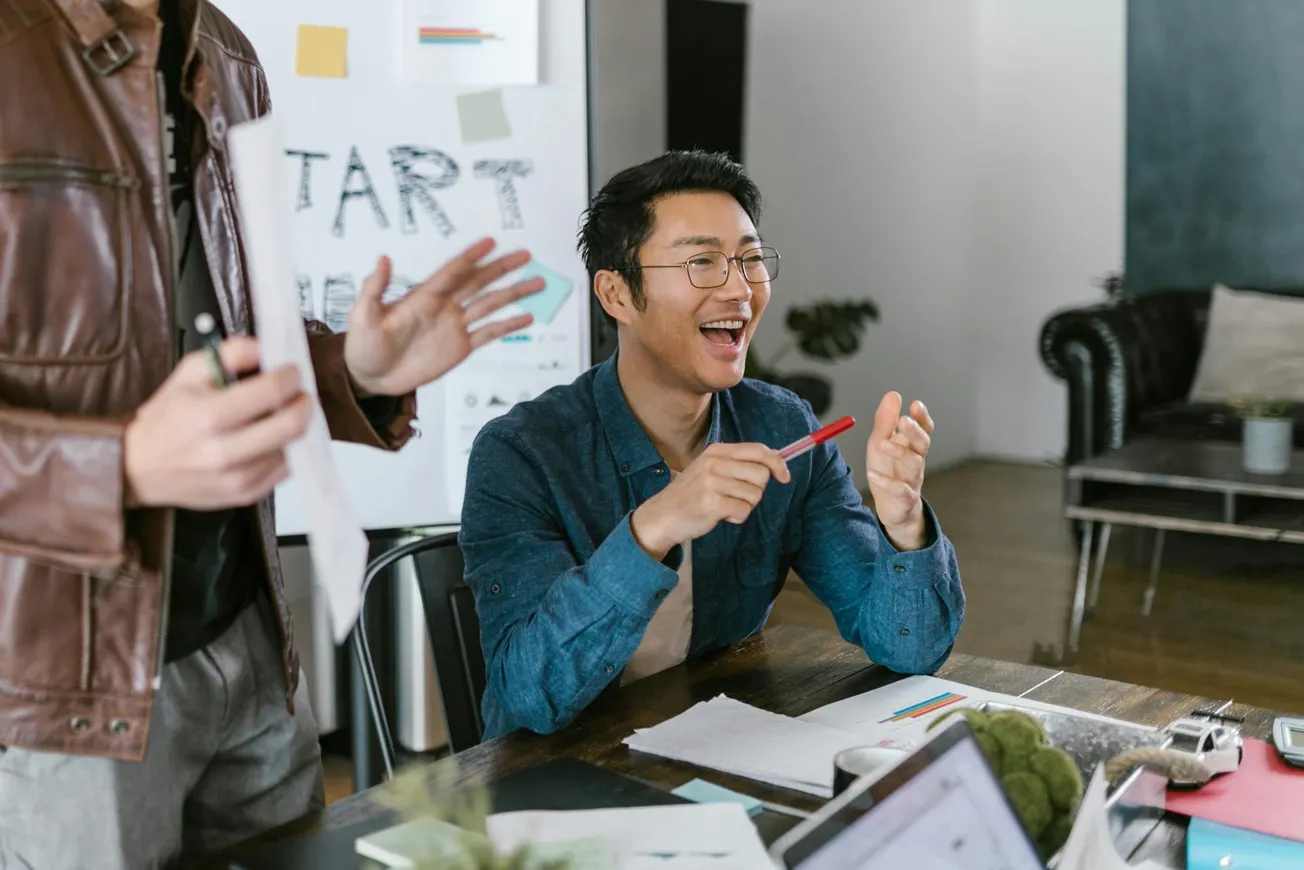 A man in glasses, holding a pen, is smiling and clapping in a bright office. A whiteboard with "Start" is visible; the atmosphere is energetic and collaborative.