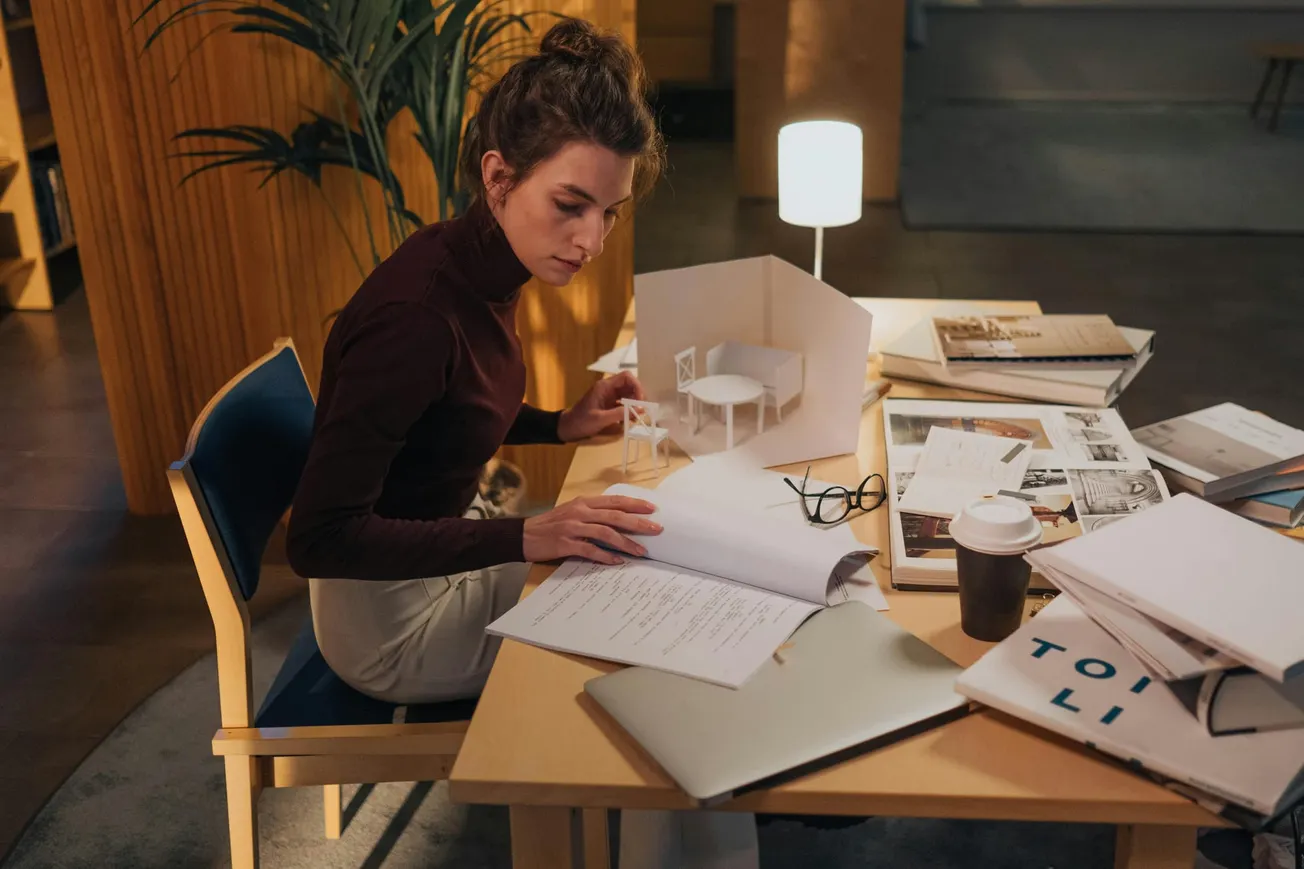 A person in a dimly-lit room focuses on architectural work at a cluttered desk, with scale models, papers, a laptop, and a coffee cup. The mood is studious.