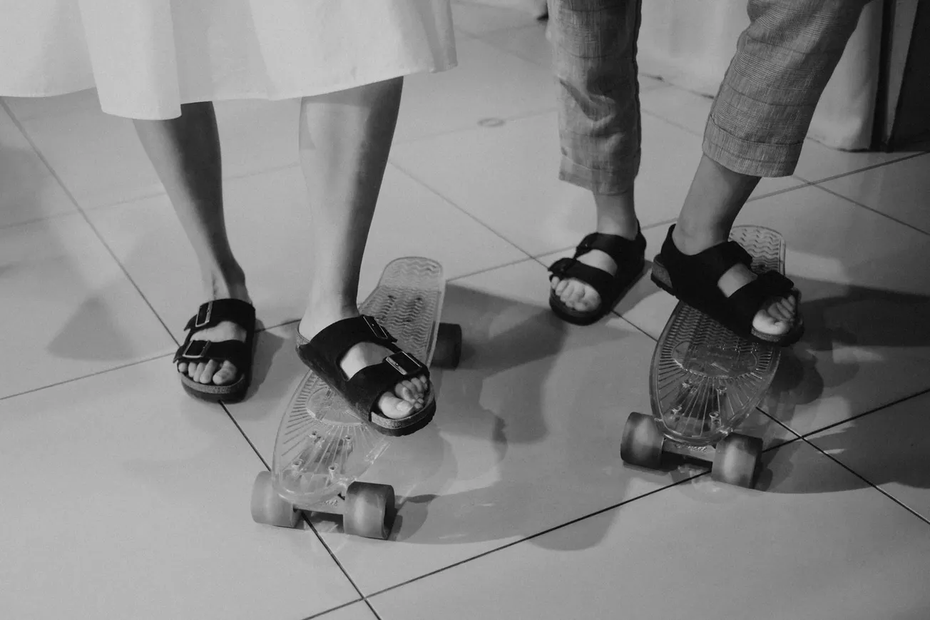 Two people stand with their feet on skateboards indoors. They're wearing sandals; one in a skirt, the other in pants. The tone is playful and casual.