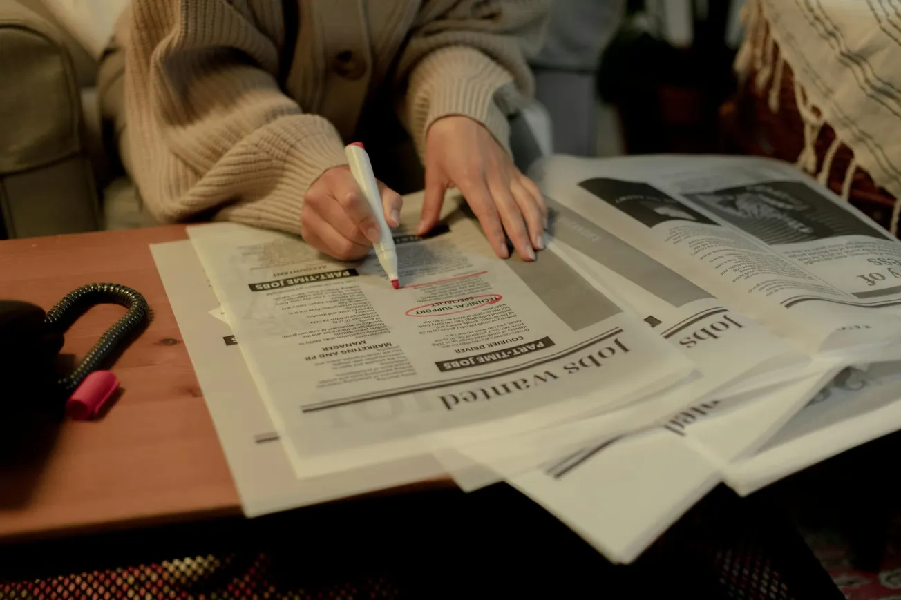 A person in a cozy sweater highlights job listings in a newspaper with a pink marker. Newspapers are spread across a wooden table, conveying focused determination.