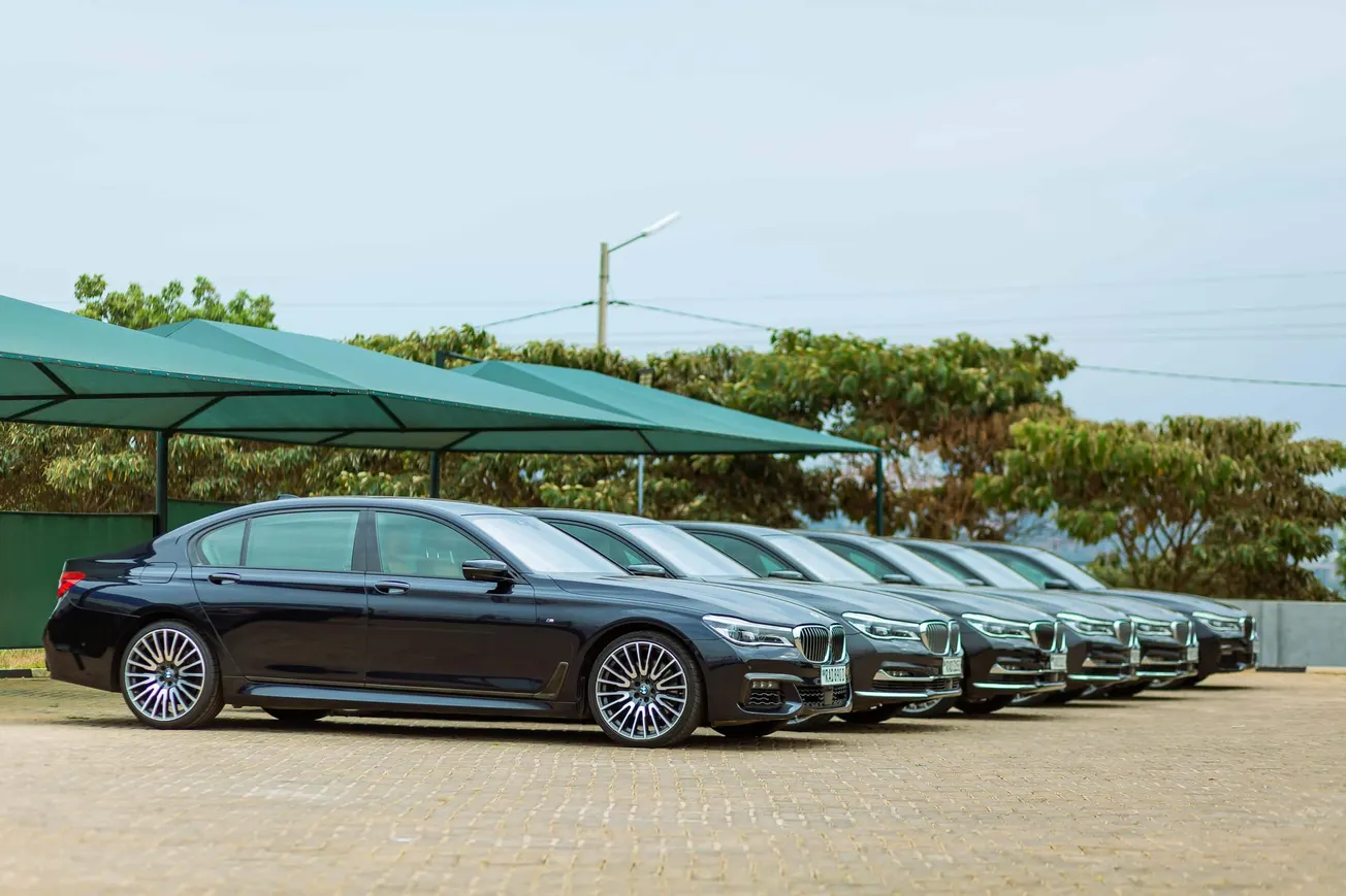 A line of six sleek black luxury sedans is parked under green canopies on a paved lot, with trees in the background under a clear sky.