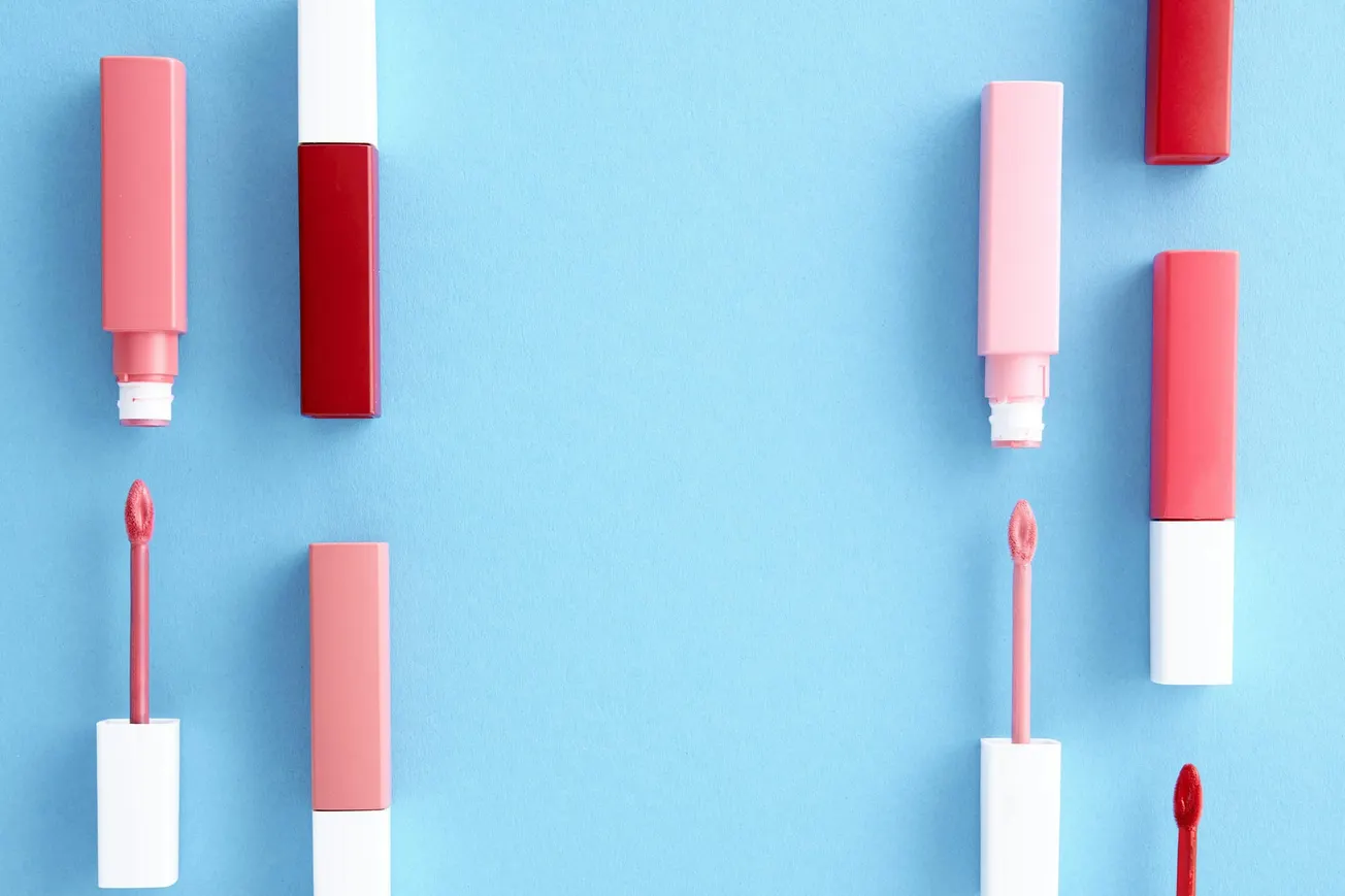 Various pink and red liquid lipsticks with open applicators are arranged on a light blue background, creating a stylish and colorful pattern.