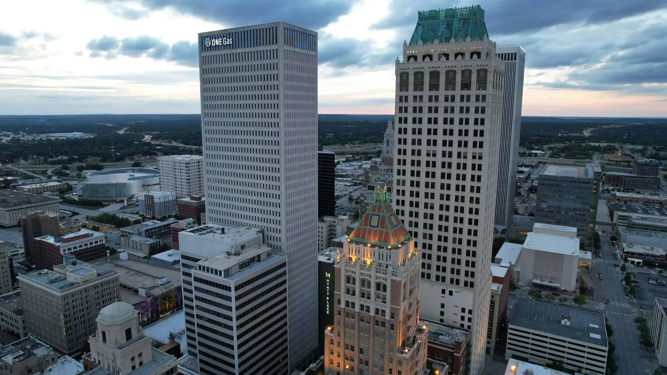 Aerial view of downtown Tulsa at dusk, with prominent skyscrapers against a cloudy sky. Buildings are lit up, creating a serene urban scene.