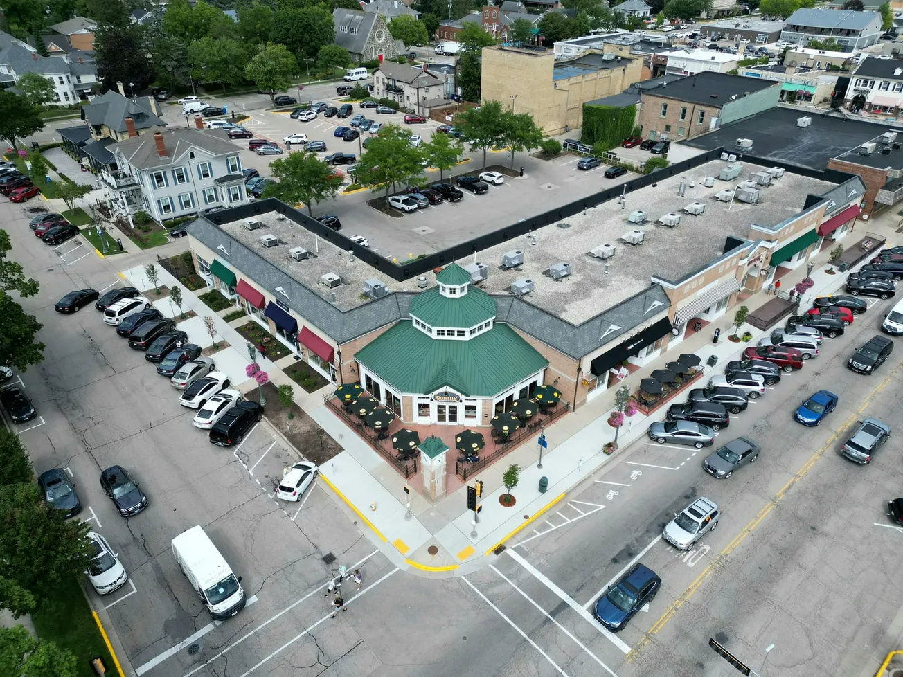 Aerial view of a bustling shopping plaza with a building featuring a green roof and outdoor seating. Cars fill parking spaces, conveying a lively atmosphere.