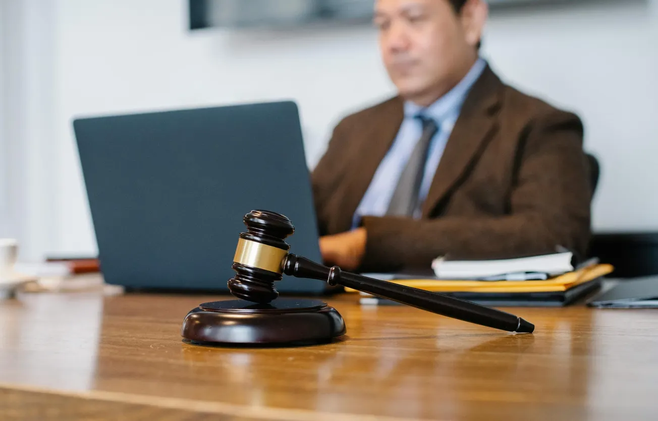 A wooden gavel rests on a table in focus, symbolizing justice. In the blurred background, a suited man works on a laptop, suggesting a legal setting.