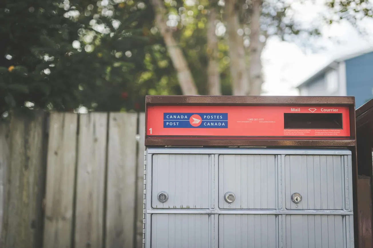 A Canada Post mailbox with a red top stands against a wooden fence, surrounded by trees in a suburban setting. The tone is calm and everyday.