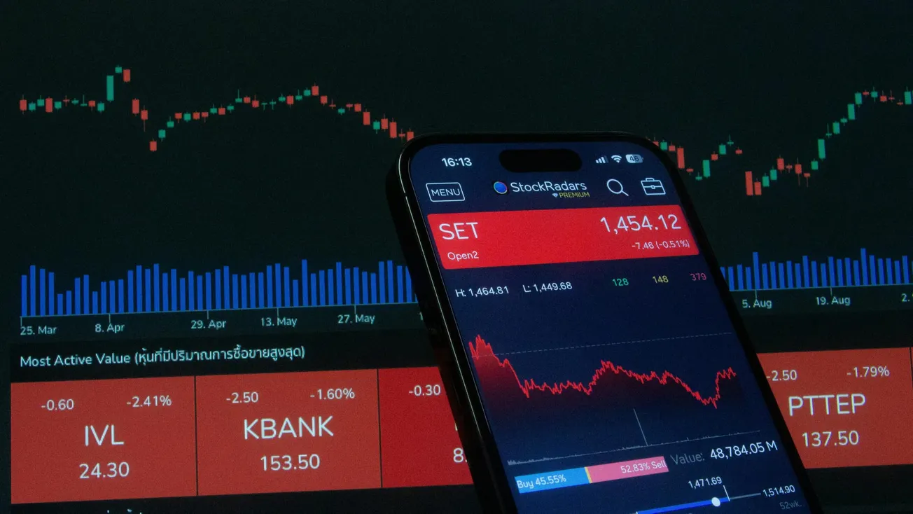 A smartphone displays a stock market app with red graphs indicating a downward trend. Behind it, a screen shows fluctuating stock charts and numbers.