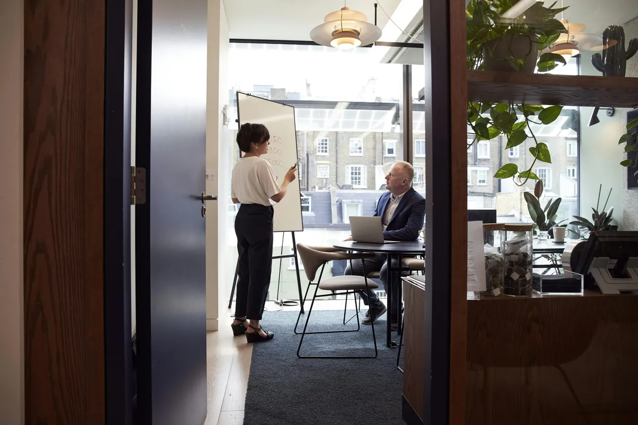 A woman presents at a whiteboard in a modern office, engaging with a seated man using a laptop. Large windows and plants create a bright, focused atmosphere.