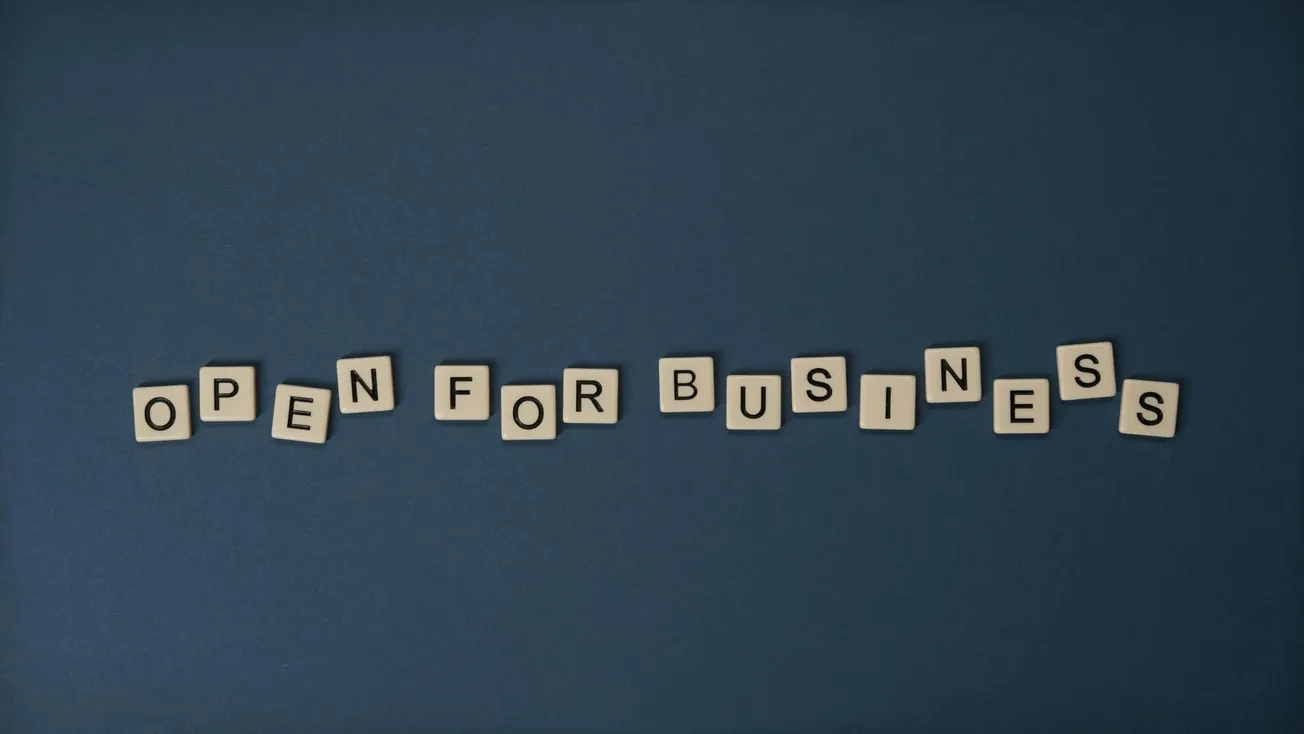 Scrabble tiles on a dark blue background spell "Open for Business." The arrangement conveys readiness and welcome, suggesting a positive, inviting tone.