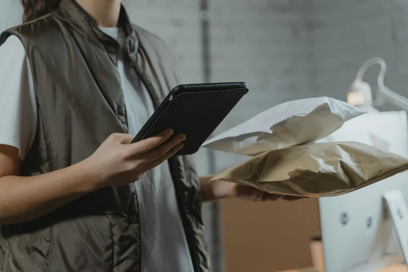 A person in a vest holds a tablet and two packages, standing near a desk in a warehouse or office environment, suggesting logistics or delivery tasks.