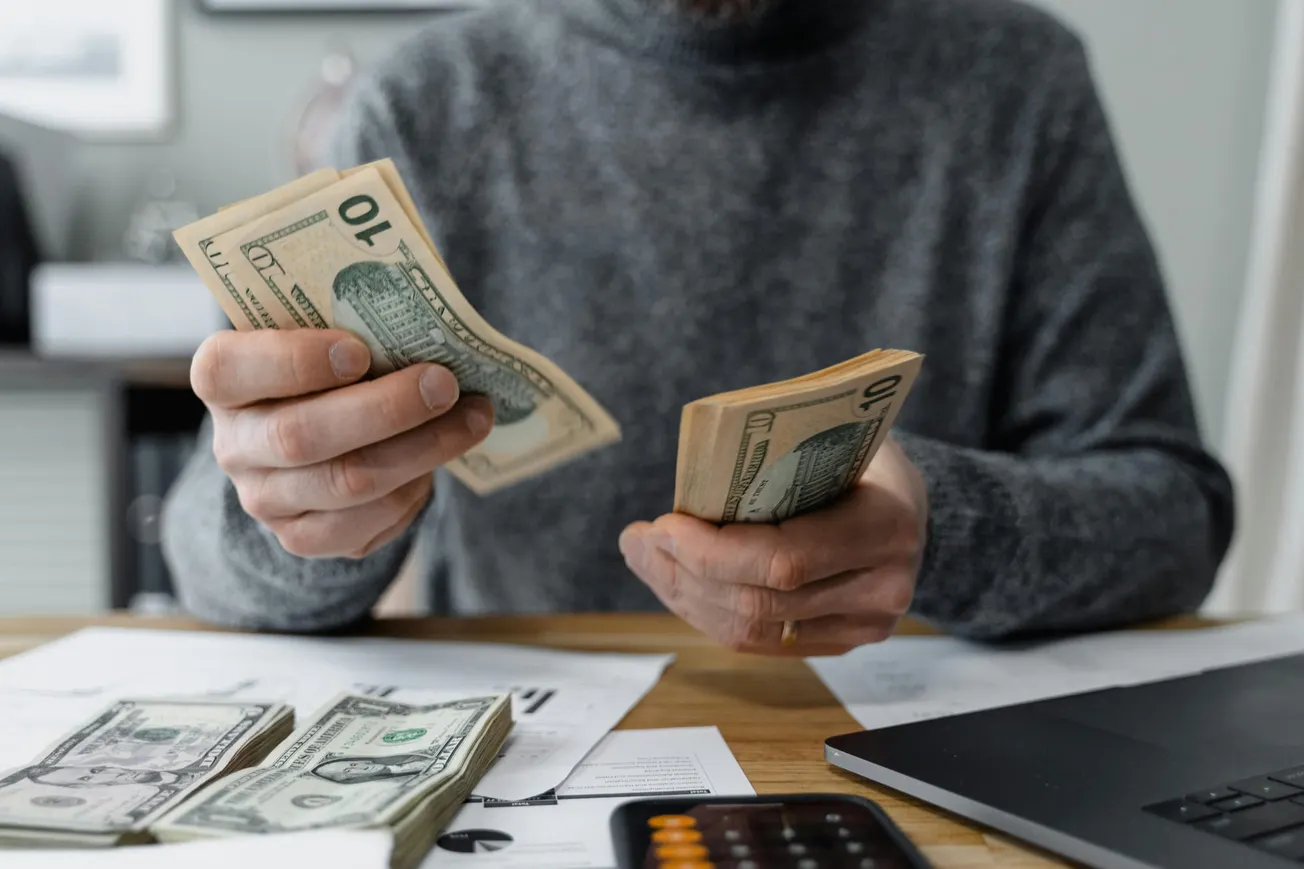 A person in a gray sweater counts stacks of U.S. dollar bills at a cluttered desk, which also has scattered papers, a calculator, and a laptop.