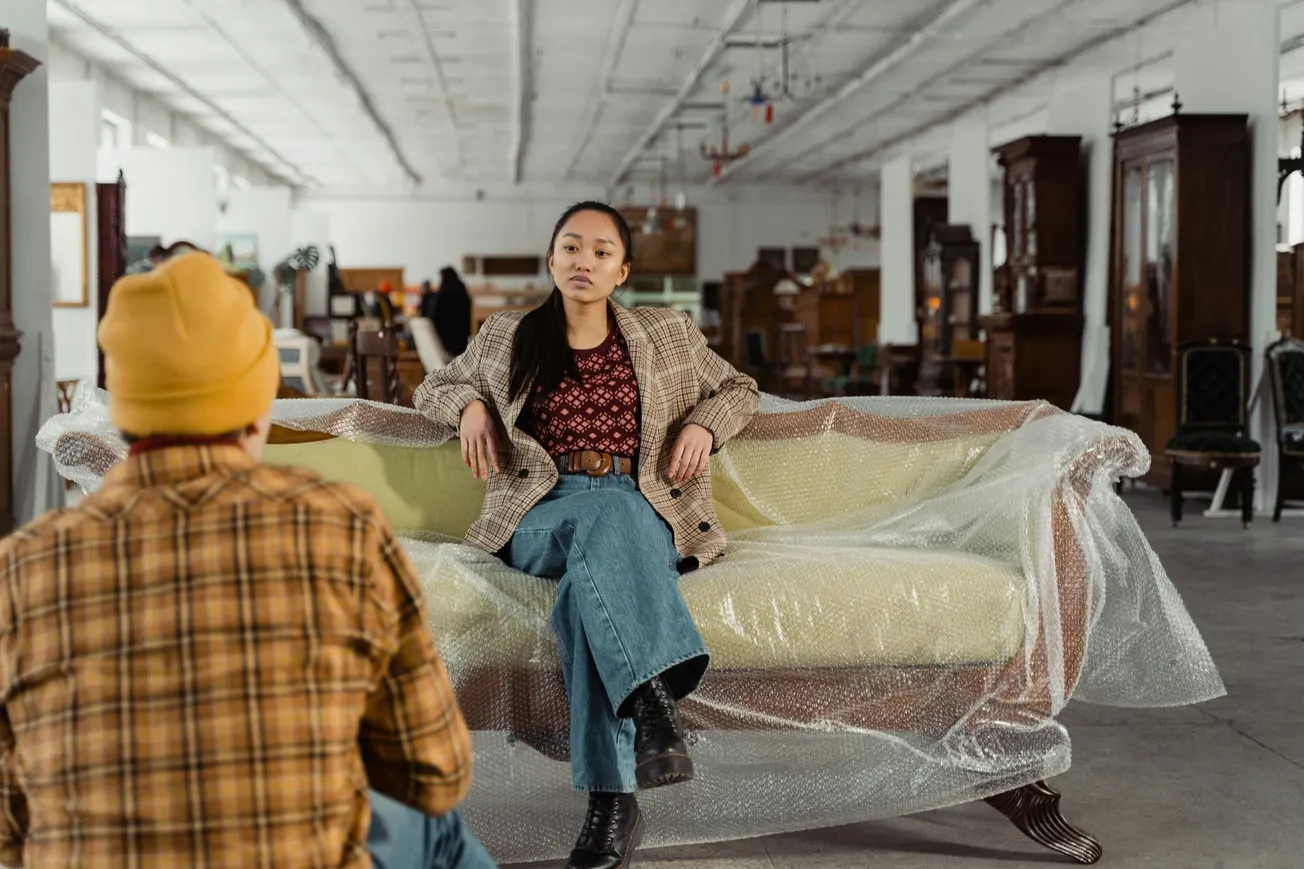 A person sits confidently on a bubble-wrapped couch in a spacious warehouse. Another person, wearing a yellow beanie and plaid shirt, faces them.