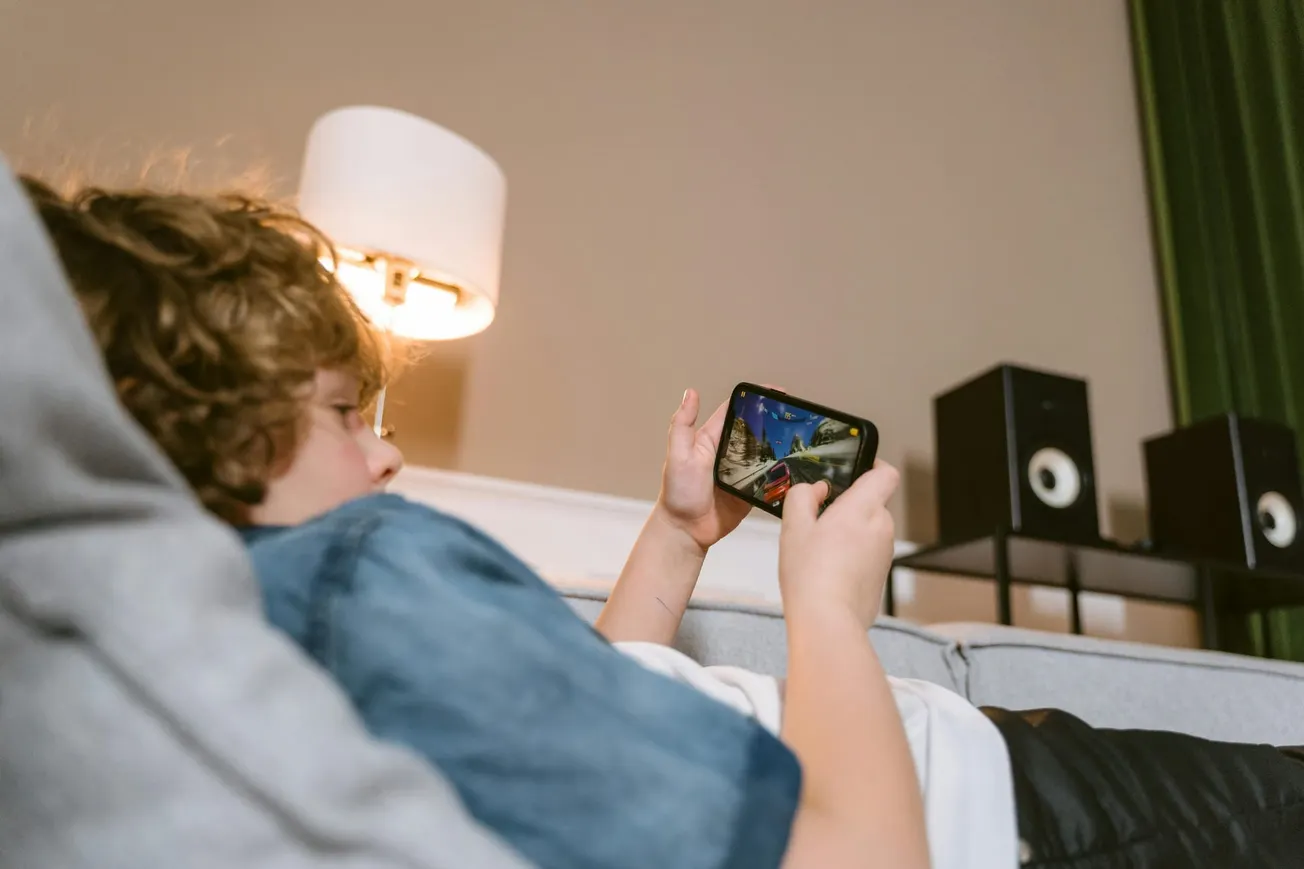 A child with curly hair, wearing a blue shirt, lies on a couch playing a video game on a smartphone. A lamp and speakers are in the background, creating a relaxed atmosphere.