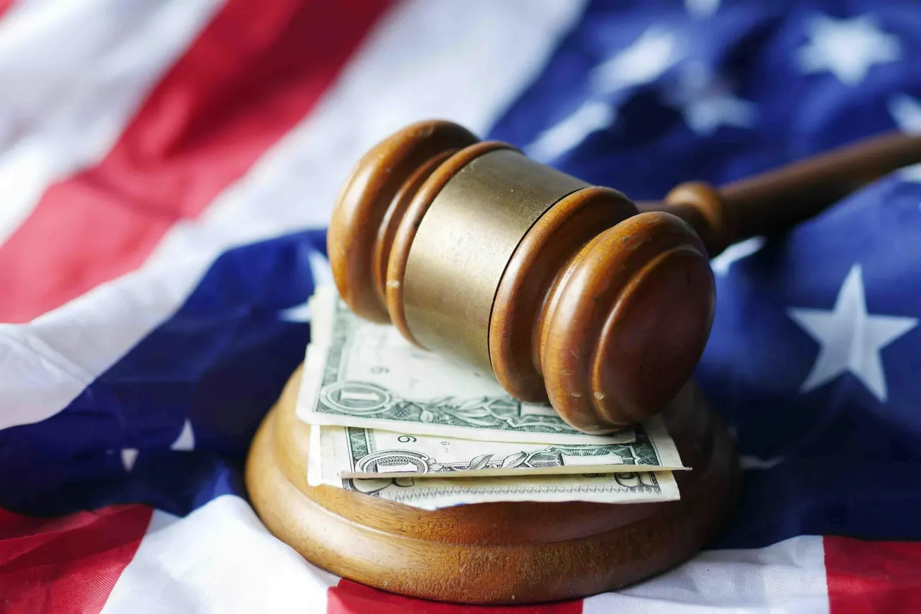 A wooden gavel rests on a stack of dollar bills, placed on an American flag. The image conveys themes of justice, law, and finance in the U.S.