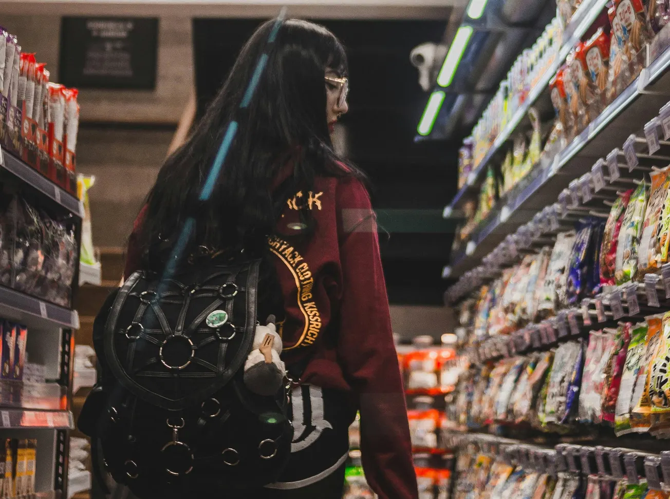 A person with long hair, wearing a red jacket and backpack, walks through a grocery store aisle lined with colorful packaged snacks.