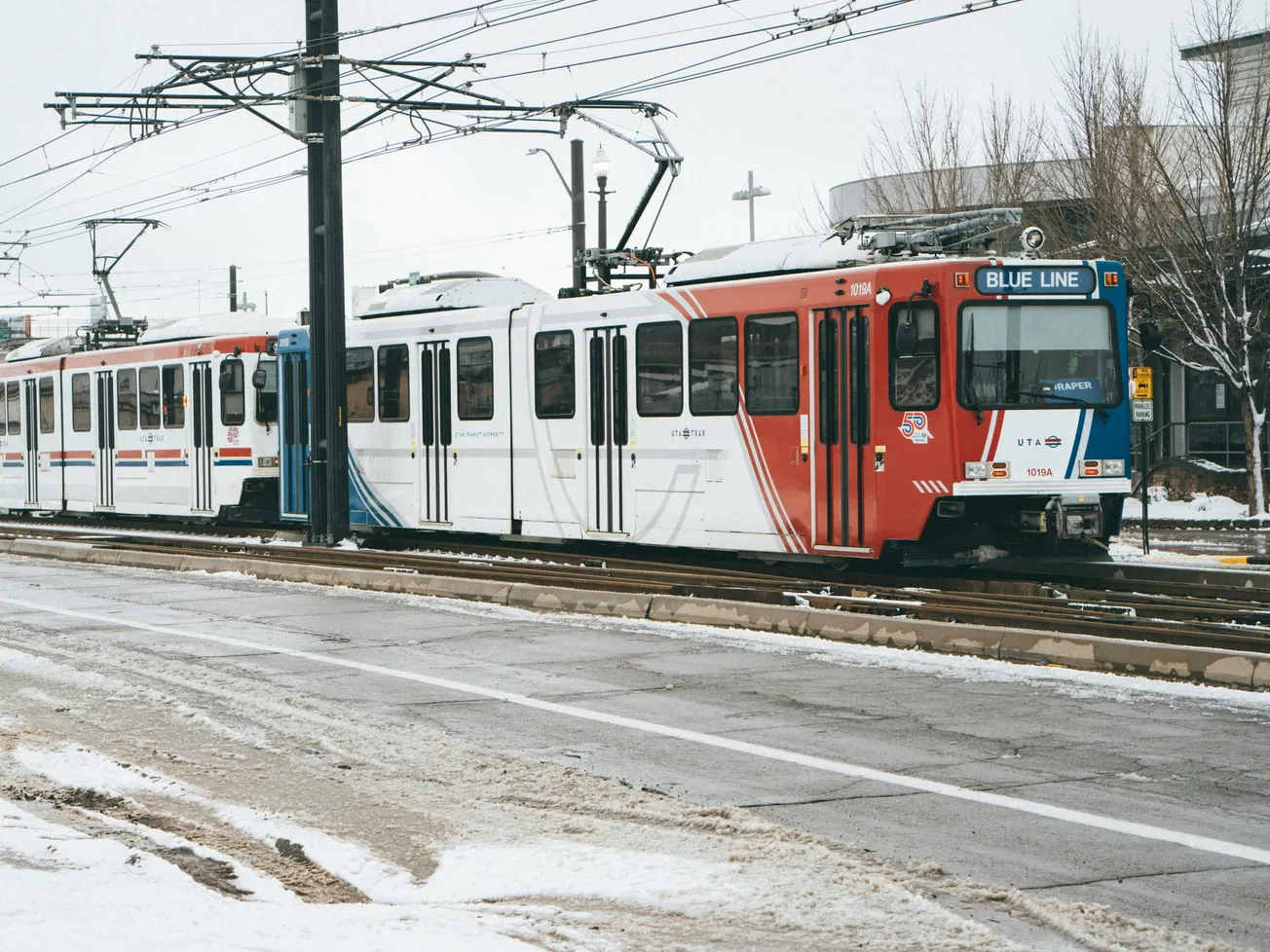 A light rail train on the snowy Blue Line track stops at a station. The sky is overcast, and bare trees surround the area, creating a wintery atmosphere.