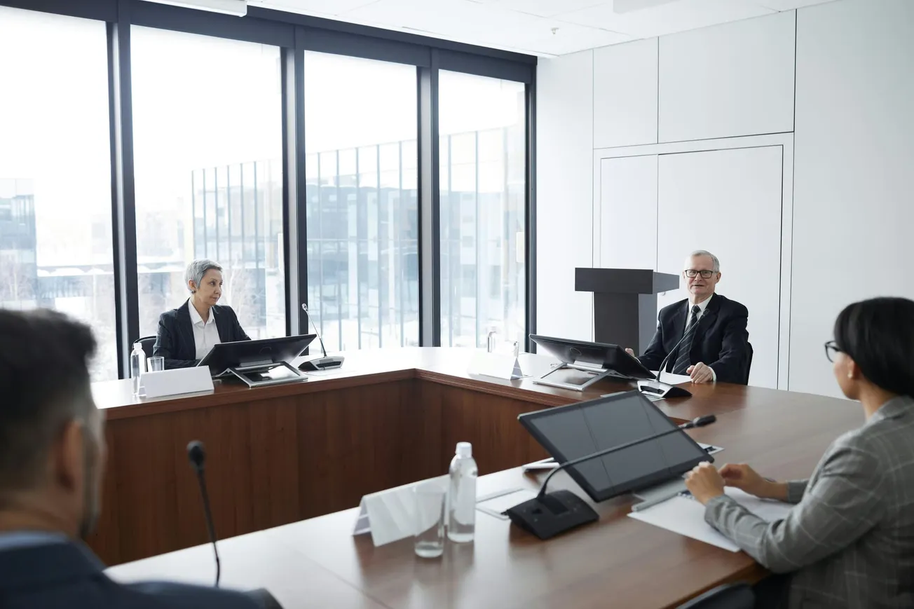 Four people in a modern conference room with large windows are engaged in a meeting. The tone is professional and collaborative.