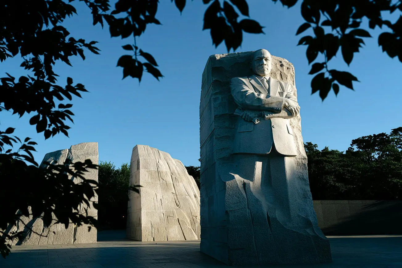 Monument of a man carved in stone stands against a clear blue sky, surrounded by large stone structures. Silhouetted leaves frame the scene.
