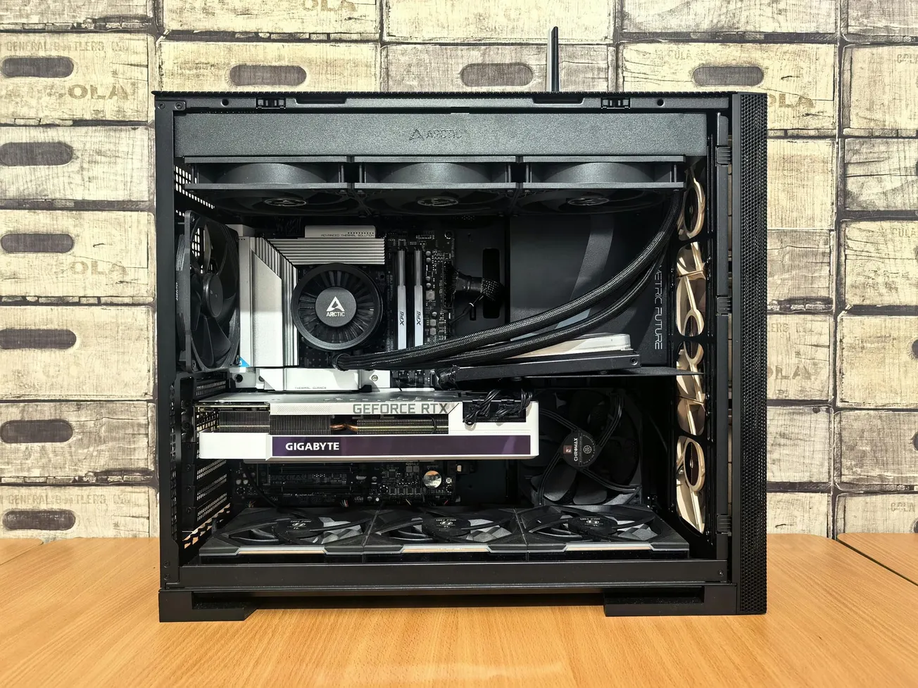 A sleek open-sided PC case revealing internal components: graphics card labeled "GeForce RTX," liquid cooling tubes, fans, and a motherboard. Wood desk, textured wall.