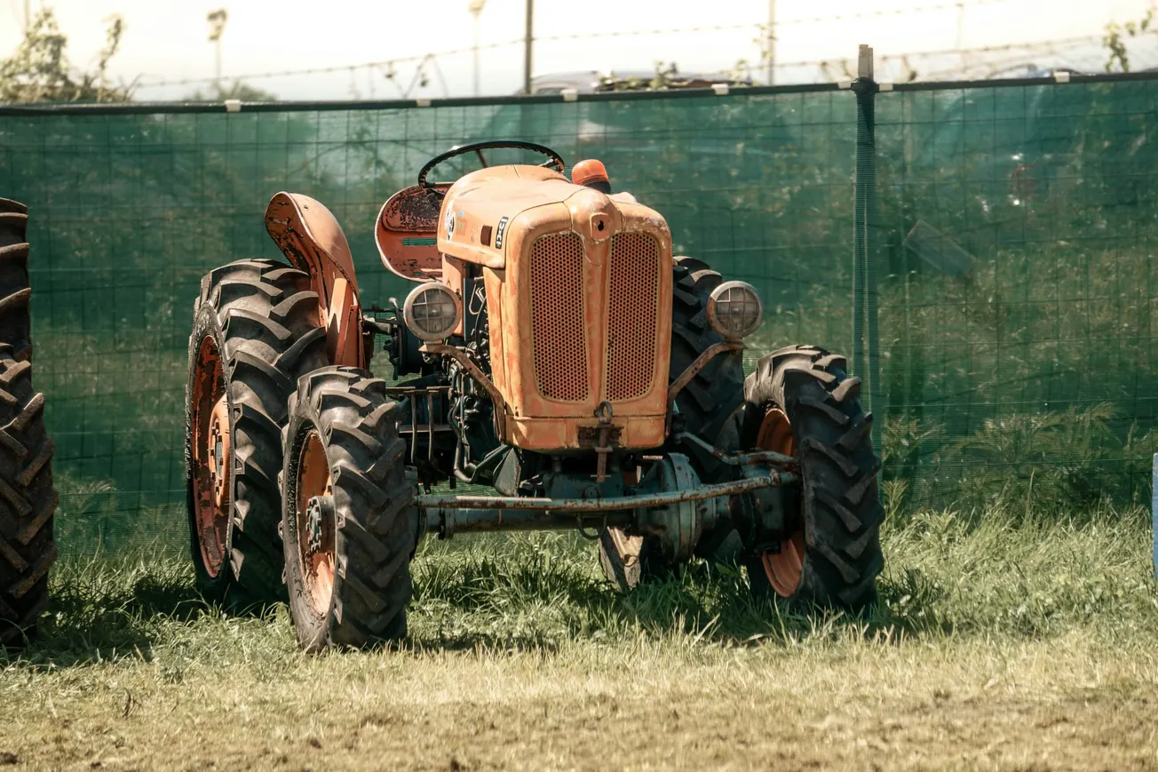 An old, rusted orange tractor with large tires sits on green grass. A green mesh fence and blurred plants in the background create a nostalgic tone.