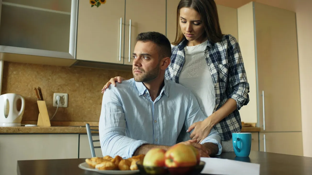 A concerned woman comforts a seated man in a kitchen, her hand on his shoulder. A plate of pastries and apples sits on the table, next to a blue mug.