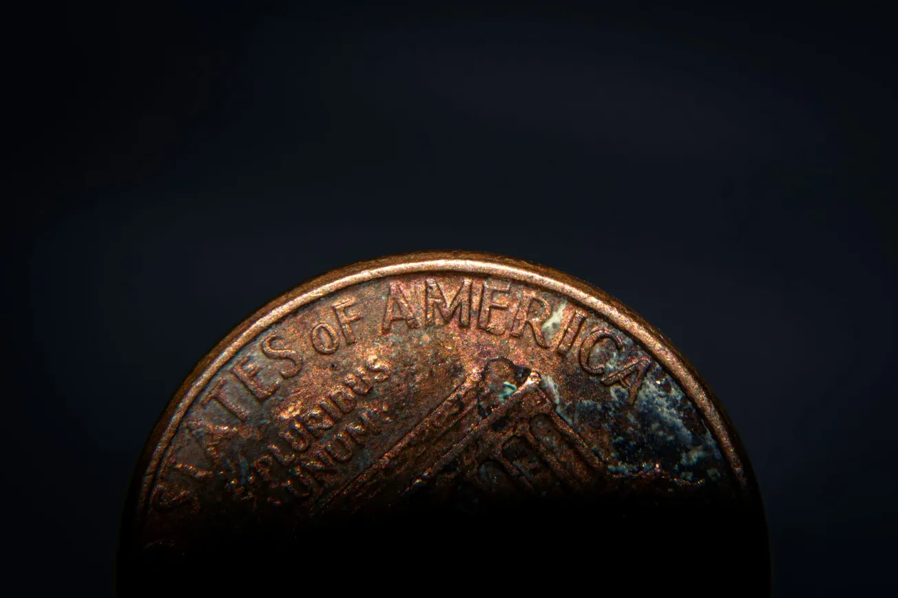 Close-up of a partially-lit, weathered US penny, with the top half visible, showing the words "States of America" against a dark background.