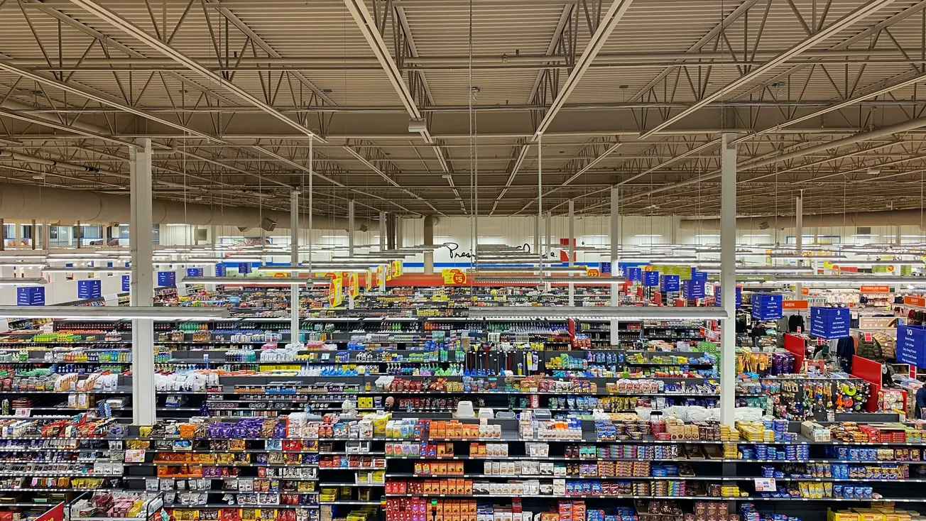 Interior of a large supermarket with neatly organized aisles featuring colorful product displays and signs under bright fluorescent lighting. The atmosphere is orderly.