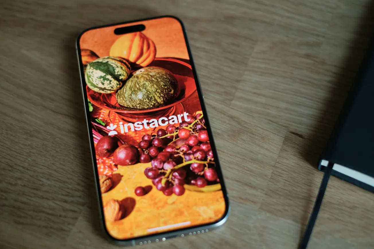 A smartphone on a wooden table displays the Instacart app with an image of assorted fruits and vegetables, conveying a fresh, vibrant feel.