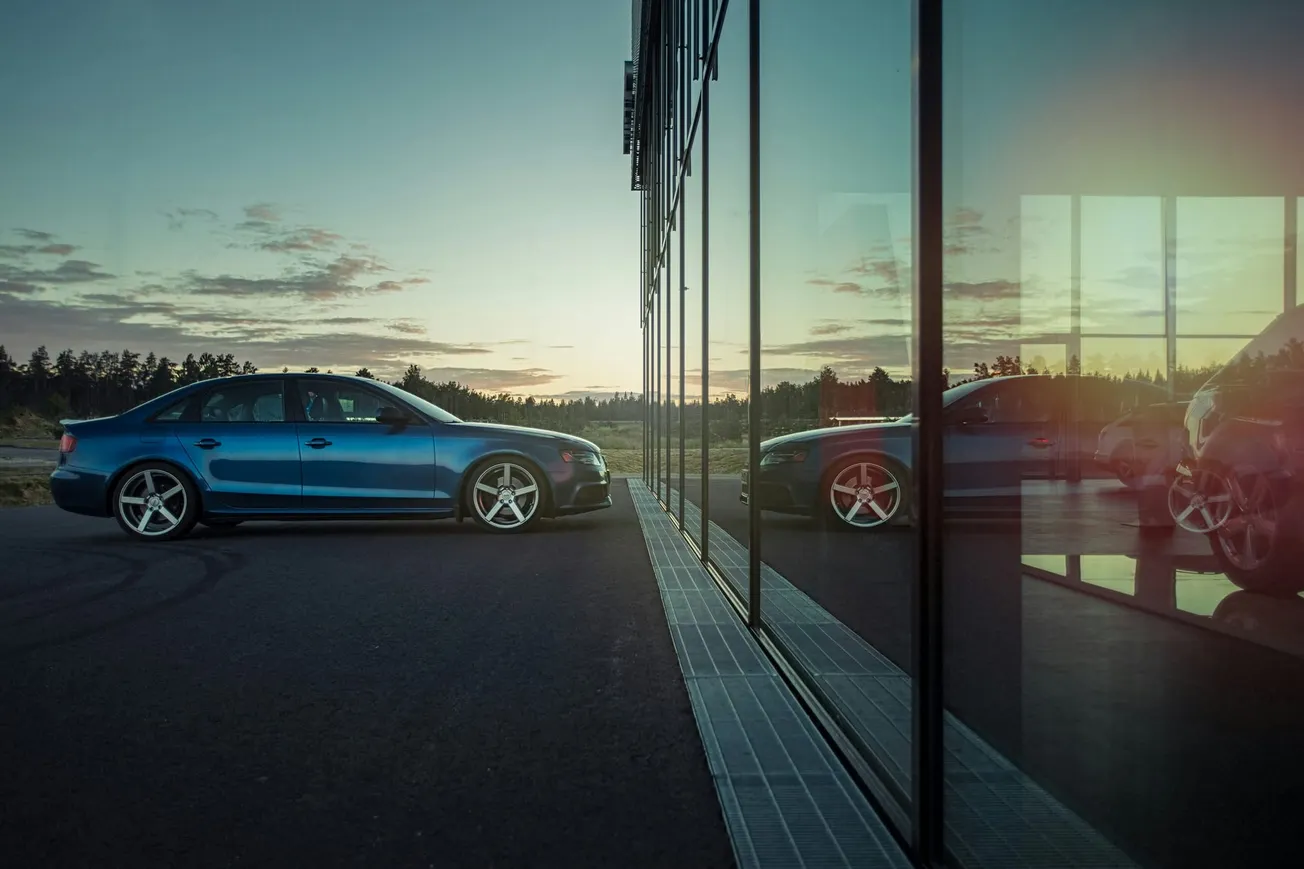 Blue car parked by a glass building at sunset, reflecting in the windows. The scene is calm, with soft light and a serene sky in the background.