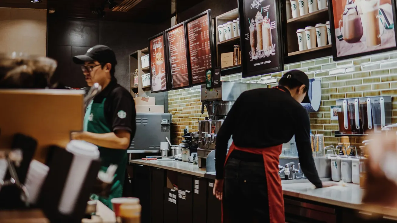 Two baristas in a busy coffee shop, wearing aprons and caps, prepare drinks. Menus and coffee cups line the wall, creating a warm, bustling ambiance.