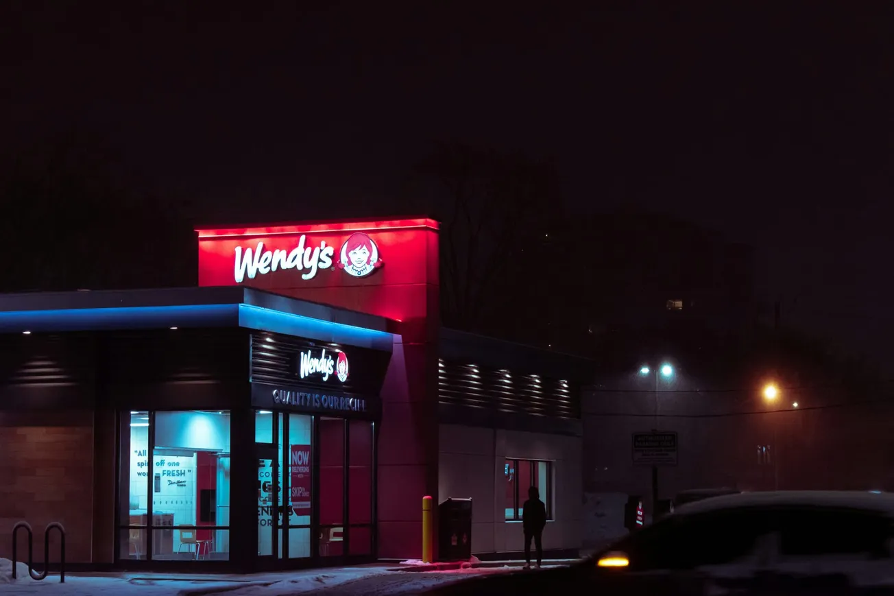 A Wendy's restaurant is lit up at night, with a glowing red sign against a dark, snowy backdrop. Few cars and a silhouette of a person create a quiet, moody atmosphere.