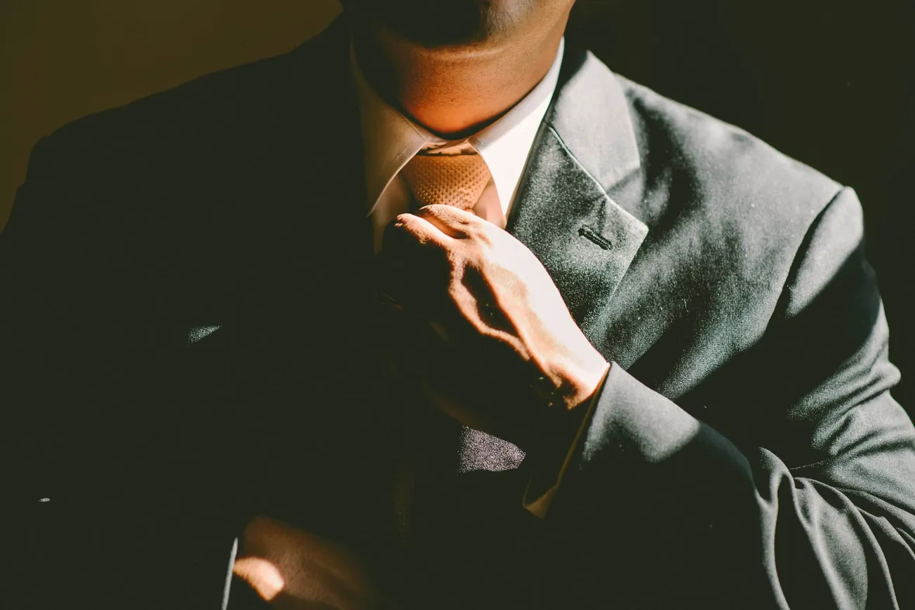A person adjusts their tie, wearing a dark suit and white shirt. Dim lighting casts dramatic shadows, conveying a professional and confident tone.