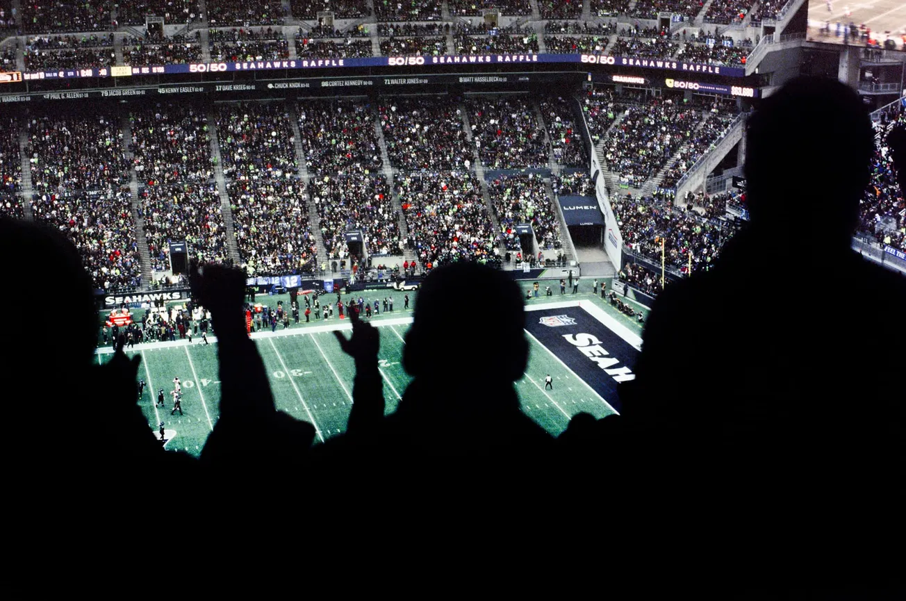 Silhouetted fans cheer in a stadium, overlooking a football field with "Seahawks" branding. The packed stands reflect a vibrant, energetic atmosphere.