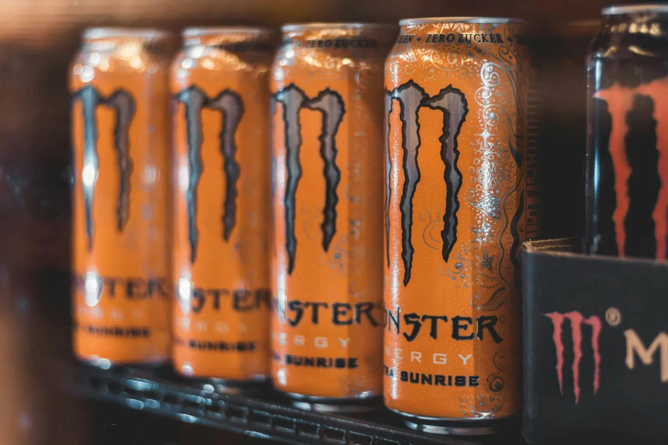 Five orange energy drink cans with a black "M" logo are arranged in a fridge. The scene conveys a bold, energetic vibe.