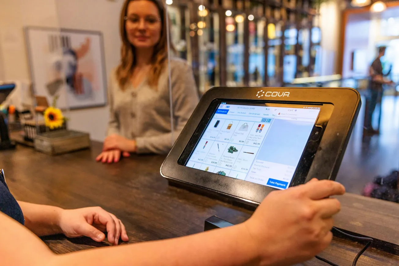 A person holds a stylus at a tablet displaying a product menu on a checkout counter. Another person stands across, smiling. Bright and welcoming atmosphere.
