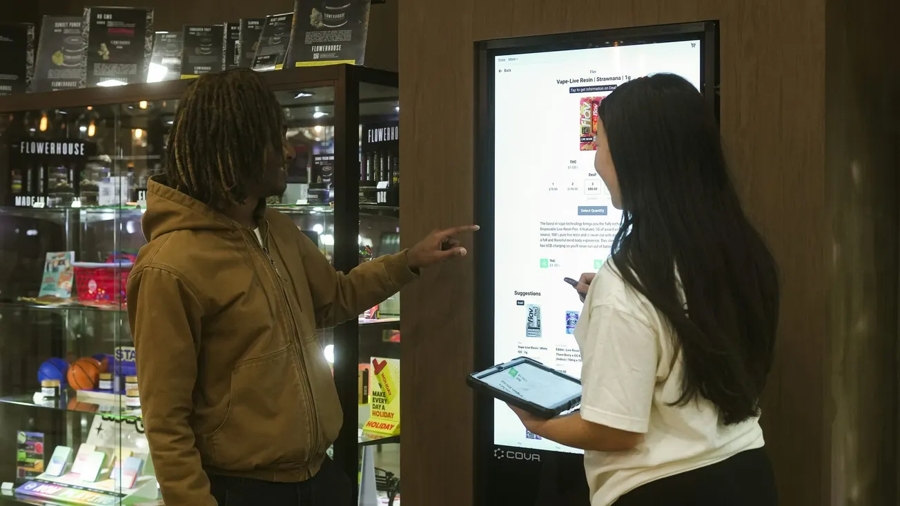Two people interact with a touchscreen kiosk in a store. One points at the screen, while the other holds a tablet. The scene conveys collaboration.