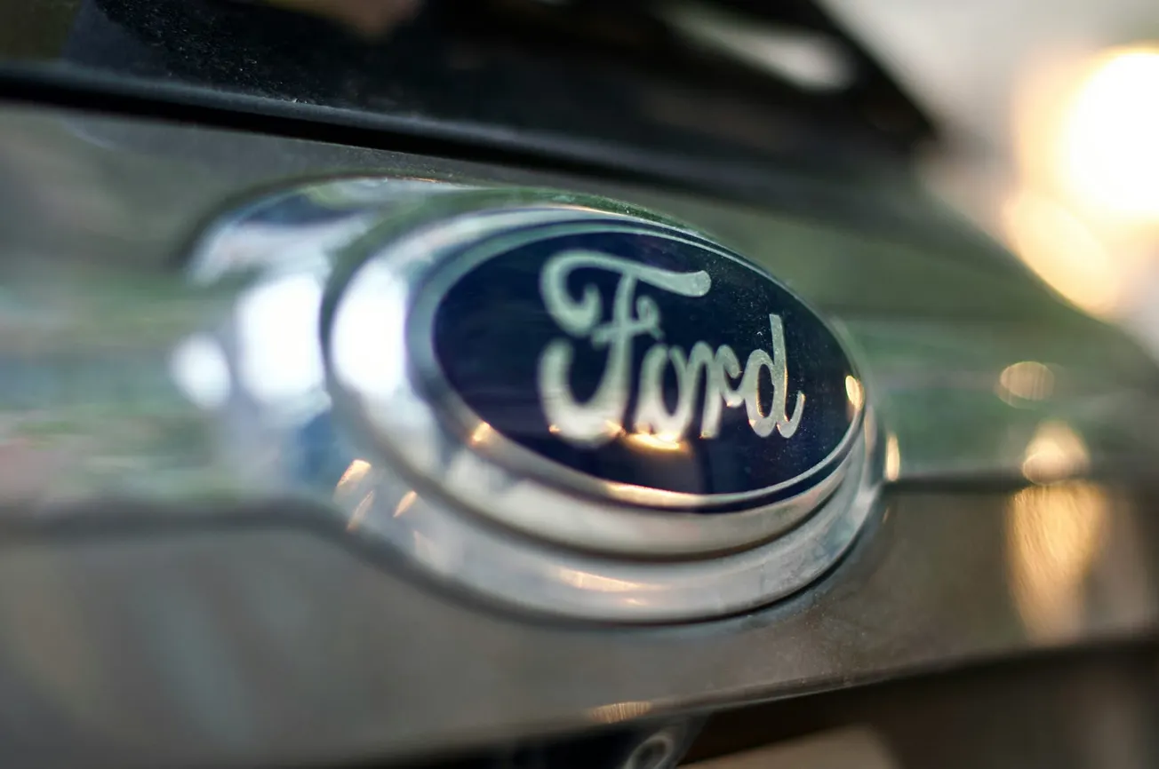 Close-up of a Ford car emblem on a vehicle grille, featuring a blue oval logo with white lettering. The background is softly blurred, creating a sleek, modern feel.