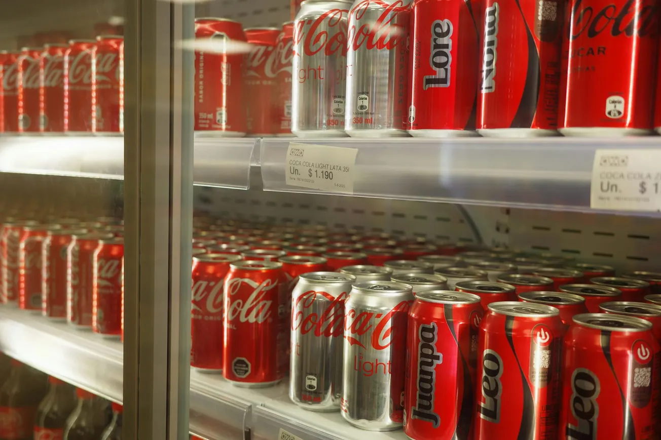 Rows of red and silver soda cans, including Coca-Cola, fill a refrigerator shelf in a store. A price tag displays 1.190 currency units.