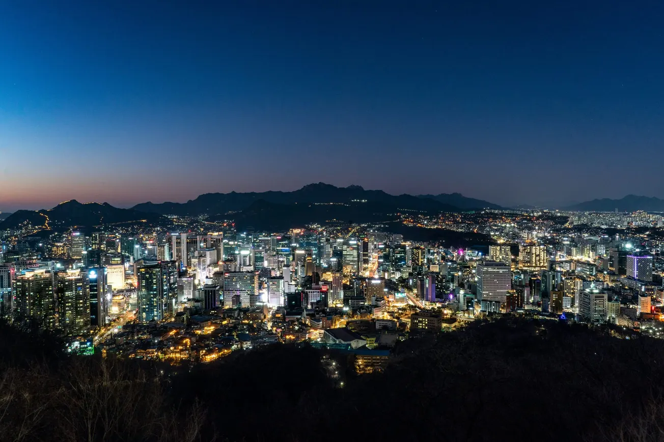 Cityscape at dusk showing a sprawling urban area densely packed with illuminated skyscrapers and buildings against a blue and fading orange sky.