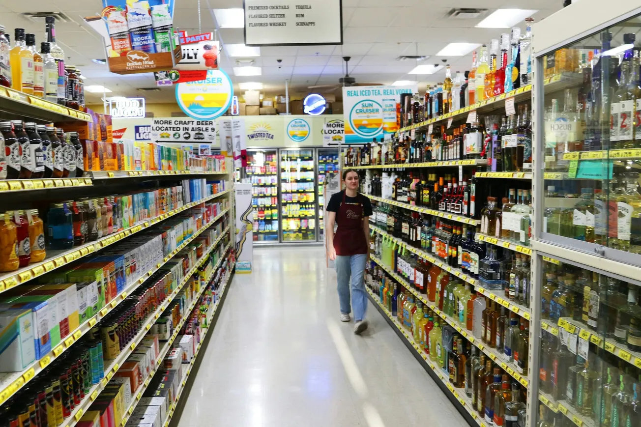 A person in a maroon apron walks down a grocery store aisle filled with colorful liquor bottles and snacks. Bright signs hang overhead, creating a busy atmosphere.