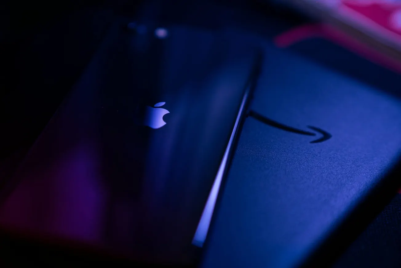 Dimly lit image showing a glossy phone with an Apple logo atop a Kindle device with an Amazon smile logo, in a moody purple and blue ambiance.