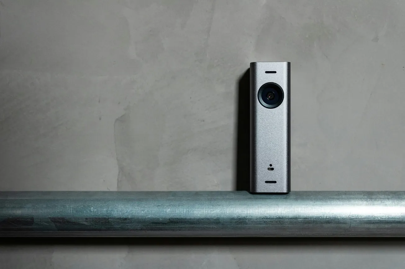 A sleek, metallic smart doorbell camera mounted on a textured gray wall above a horizontal metal pipe, casting a small shadow, evoking a modern and secure feel.