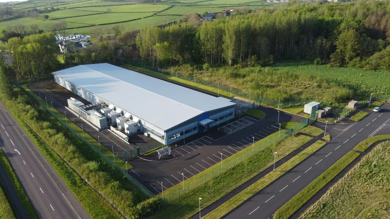 Aerial view of a large industrial building with a gray roof, surrounded by lush greenery and trees. Empty parking lot and roads are visible. Natural, serene setting.