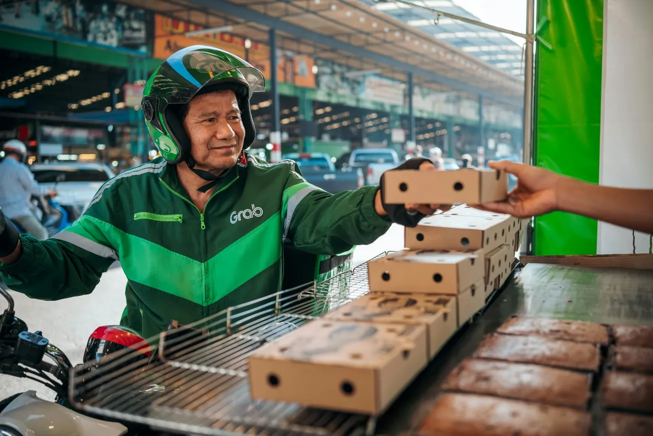 A delivery driver in a green uniform and helmet is joyfully receiving food boxes from a vendor. Background shows a bustling street market.