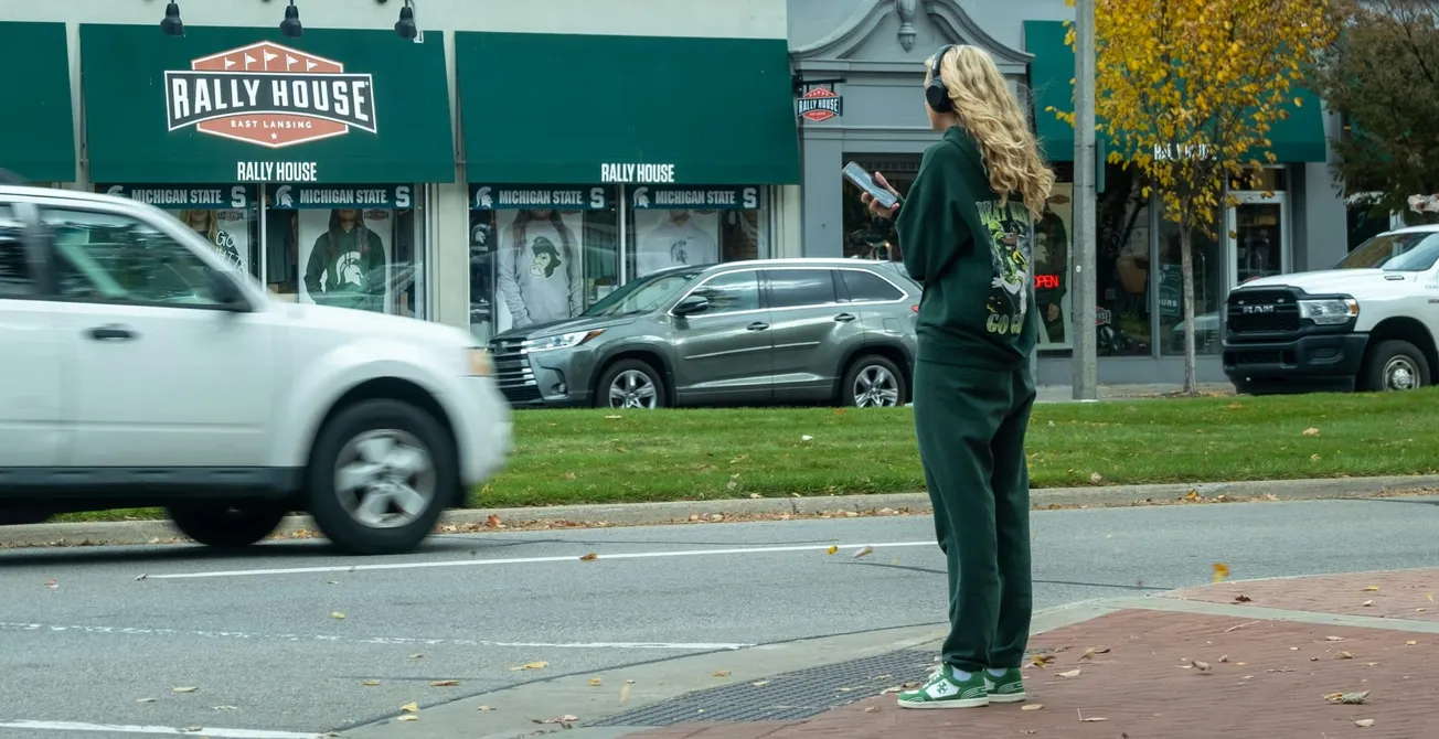 A person with long blonde hair wears a green tracksuit and sneakers, stands on a city sidewalk, holding a phone. Cars drive by, and a store called "Rally House" is visible across the street.