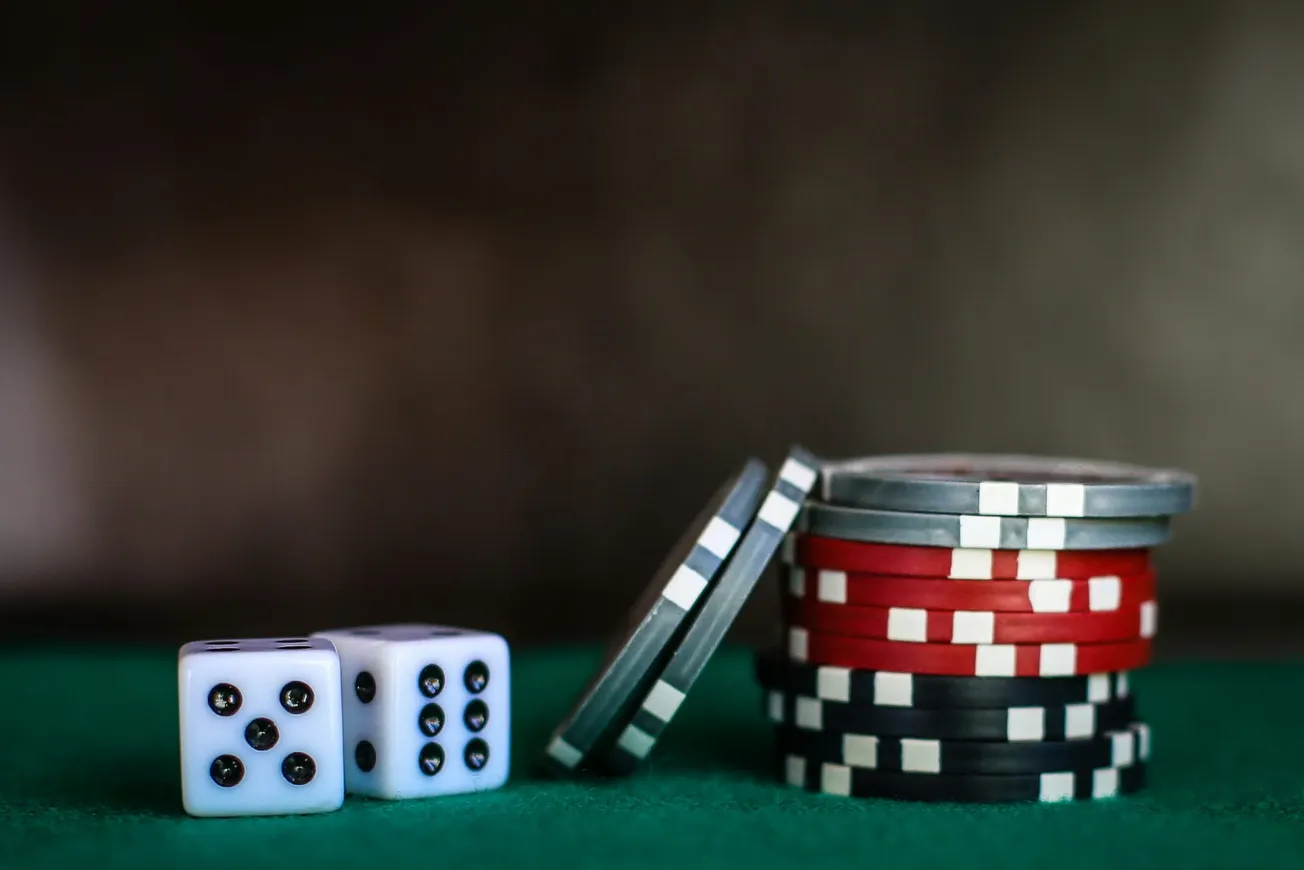 Two white dice showing four and three sit on green felt beside a stack of poker chips in red, blue, and black. The scene conveys a gambling theme.