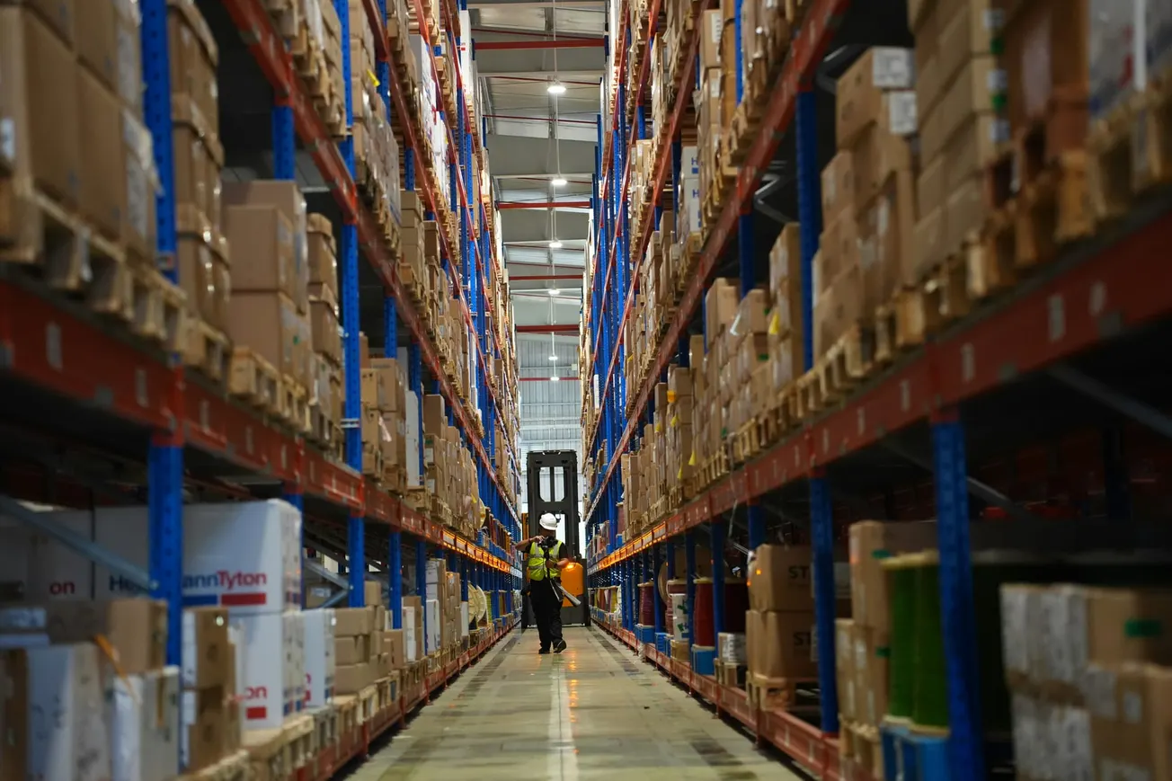 A vast warehouse aisle lined with tall shelves filled with boxes. A worker in a safety vest and helmet walks beside a forklift, conveying industriousness.