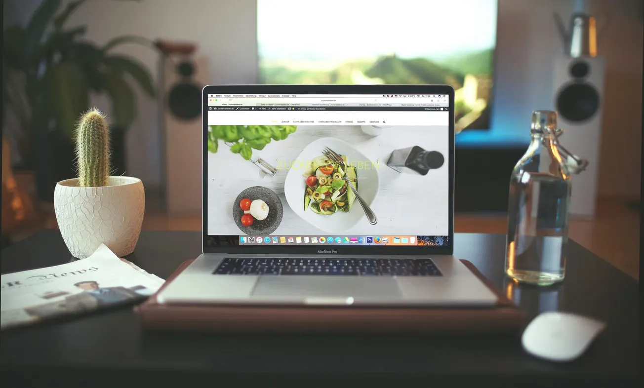 A laptop on a desk displays a website with a salad image. Nearby are a cactus, water bottle, newspaper, and mouse. The scene feels modern and cozy.