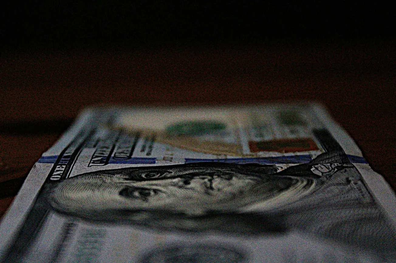 A close-up of a U.S. hundred-dollar bill on a dark surface. The focus is on Benjamin Franklin's portrait, with blurred, moody lighting conveying a somber tone.